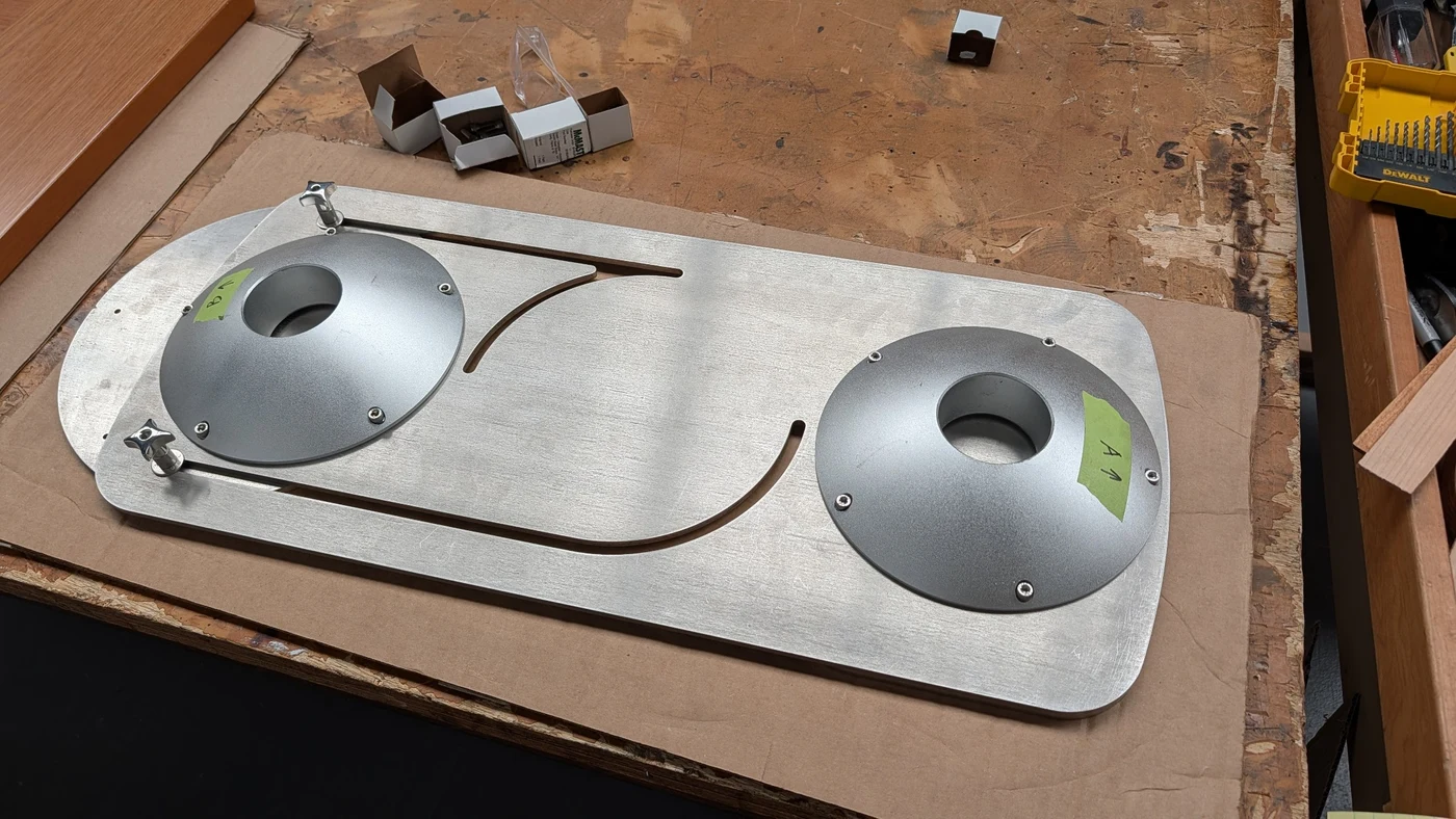 Laser-cut stainless-steel plate with curved tracks, mounting plates, and two clamping knobs on the workshop bench.